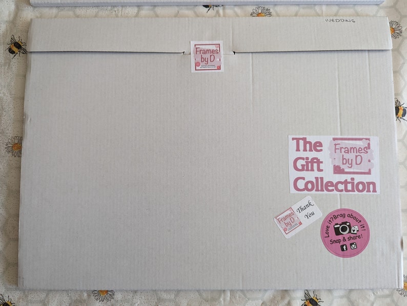 May include: A large, rectangular cardboard package with the text "WEDDING" printed on the top flap. The package features several stickers, including "Frames by D" and "The Gift Collection". A pink sticker encourages sharing on social media.
