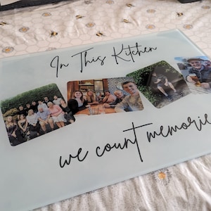 May include: A rectangular glass cutting board with the text "In This Kitchen we count memories" in black script. The board features three photos of people. The background is a white and yellow floral pattern.