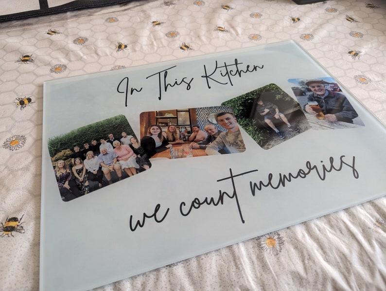 May include: A rectangular glass cutting board with the text "In This Kitchen we count memories" in black script. The board features three photos of people. The background is a white and yellow floral pattern.