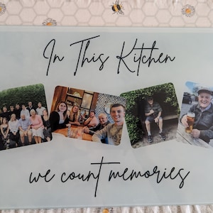May include: A rectangular glass cutting board with the text "In This Kitchen we count memories" in black script. The board features four photo collages of people. The background is a light, frosted glass.