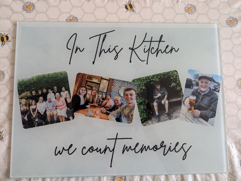 May include: A rectangular glass cutting board with the text "In This Kitchen we count memories" in black script. The board features four photo collages of people. The background is a light, frosted glass.