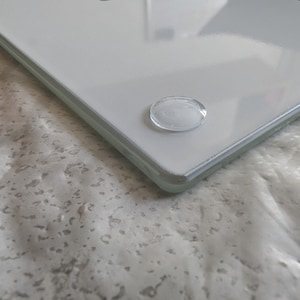 May include: A clear, rectangular glass cutting board with rounded corners. The board has a white surface and is elevated by four small, clear, oval-shaped feet. The glass reflects light, creating highlights and shadows.