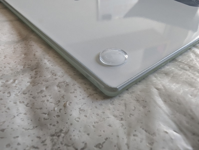 May include: A clear, rectangular glass cutting board with rounded corners. The board has a white surface and is elevated by four small, clear, oval-shaped feet. The glass reflects light, creating highlights and shadows.