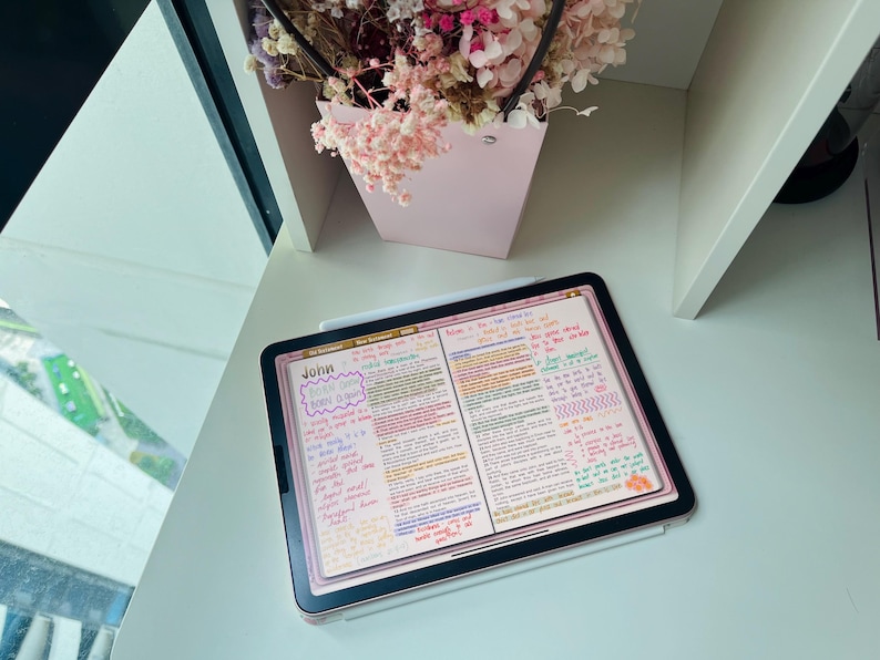 May include: A digital tablet with a pink background and handwritten notes in various colours. The text "John" is visible in the top left corner of the screen. The tablet is on a white surface with a pink flower arrangement in the background.