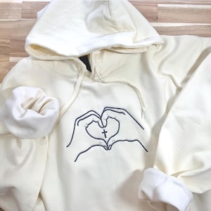 Op de afbeelding: Crèmekleurige hoodie met een capuchon met trekkoord en lange mouwen. De voorkant heeft een zwarte omtrek van twee handen die een hart vormen, met een kruis in het midden. De manchetten zijn opgerold.