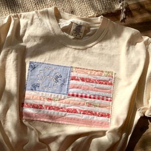 Op de afbeelding: Crèmekleurig T-shirt met een opgenaaide Amerikaanse vlagpatch. De vlag heeft een blauw veld met geborduurde sterren en het jaartal "1776", en rode en roze strepen. Het shirt is van het merk Comfort Colors, maat L-G.