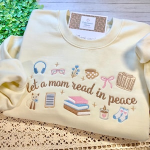 May include: Cream-colored sweatshirt with embroidered illustrations of reading-related items, including books, glasses, and a cat. The text "let a mom read in peace" is embroidered on the front. A small card is attached to the neckline.