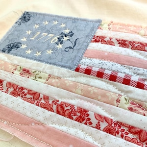 May include: A handmade textile American flag with a light blue square featuring the year "1776" and stars. The flag's stripes are made of various patterned fabrics in red, pink, and white, creating a patchwork design. The flag is on a cream-colored background.