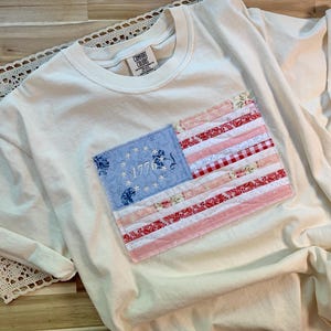 May include: Cream-coloured t-shirt with a sewn-on American flag patch. The flag features a blue field with embroidered stars and the year 1776, along with red and white striped fabric. The shirt has rolled-up sleeves.