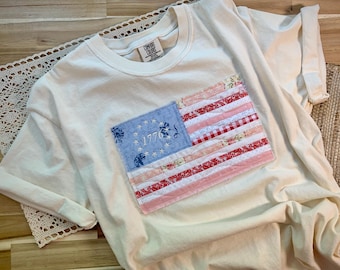 May include: Cream-colored t-shirt with a sewn-on American flag patch. The flag features a blue field with embroidered stars and the year 1776, along with red and white striped fabric. The shirt has rolled-up sleeves.