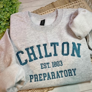 May include: A light gray sweatshirt with the word "CHILTON" embroidered in teal block letters. Below the name are the words "EST. 1803" and "PREPARATORY" in teal. The sweatshirt is folded on a textured surface.