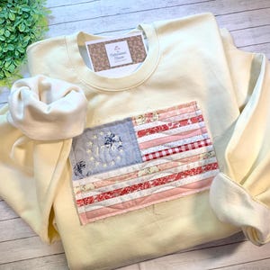 May include: A pale yellow sweatshirt with a sewn-on American flag patch. The flag features red and white stripes, a blue field with stars, and the number 1776. The sweatshirt has a crew neck and long sleeves.