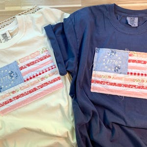 May include: Two t-shirts, one cream and one navy blue, each featuring a fabric American flag patch. The flag patches have a blue field with white stars and the year "1776", and red and white striped sections. The shirts are casual wear.
