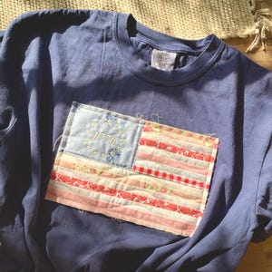 May include: A navy blue t-shirt with a sewn-on patch depicting a vintage American flag. The flag patch features red and white stripes, a blue field with embroidered stars, and the year "1776". The shirt is made of soft material.