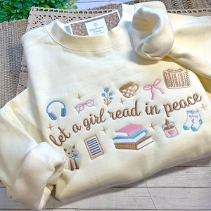 May include: Cream-colored sweatshirt with embroidered design. The design includes the text "Let a girl read in peace" along with illustrations of books, headphones, glasses, a cat, a cup, a bow, a sweater, a candle, and socks.
