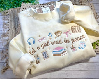 May include: Cream-colored sweatshirt with embroidered design. The design includes the text "Let a girl read in peace" along with illustrations of books, headphones, glasses, a cat, a cup, a bow, a sweater, a candle, and socks.