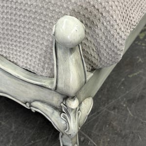 May include: Close-up of a light gray upholstered furniture piece. The furniture features ornate, carved details and a rounded top. The fabric is a textured, light gray material. The legs are painted in a distressed, light gray finish.