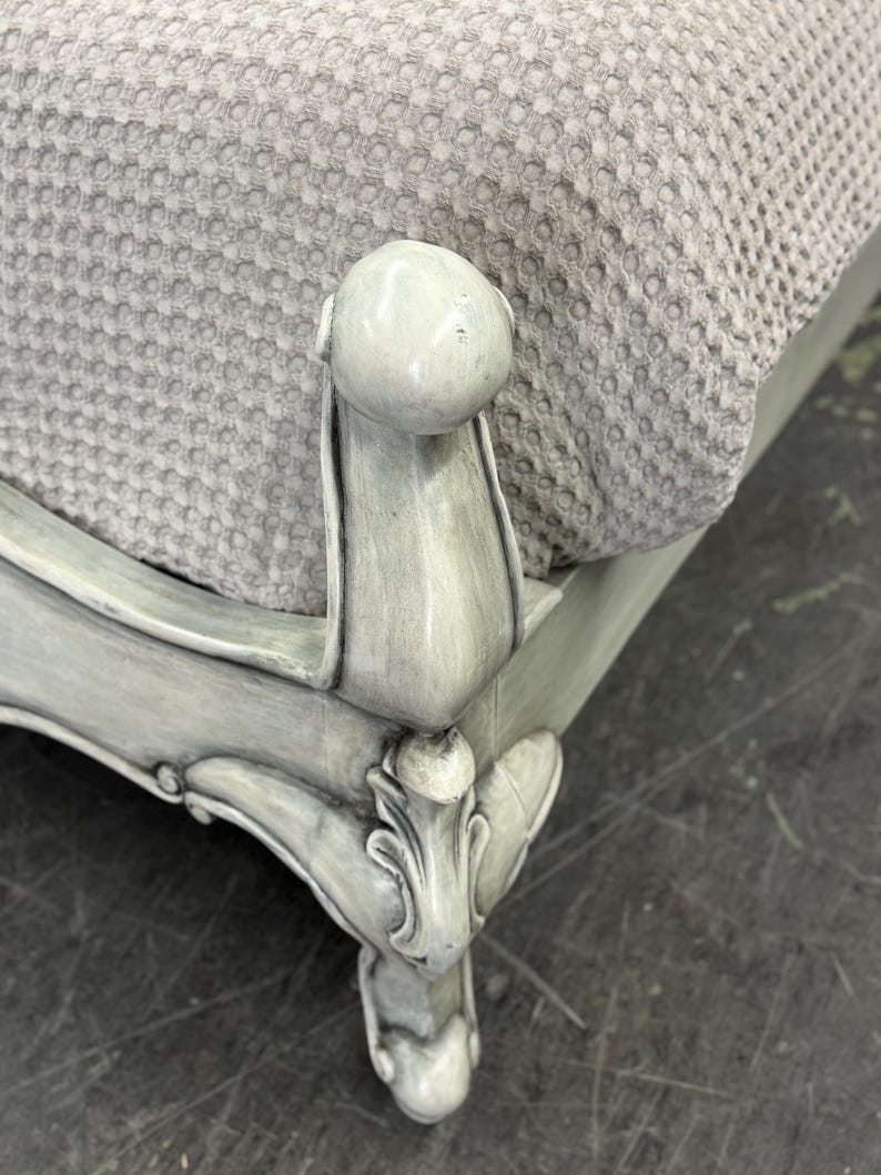 May include: Close-up of a light gray upholstered furniture piece. The furniture features ornate, carved details and a rounded top. The fabric is a textured, light gray material. The legs are painted in a distressed, light gray finish.