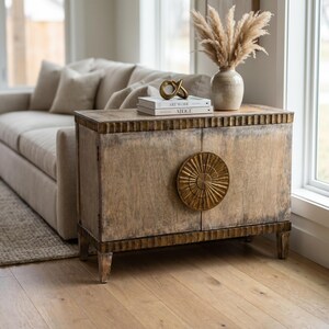 May include: A rectangular wooden cabinet with a distressed light brown finish, featuring two doors with a large, circular, gold-toned sunburst-style metal handle. The cabinet has a fluted trim along the top and bottom edges and four tapered legs.