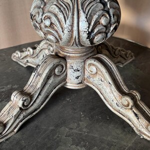 May include: A white and grey distressed wooden table base with a three-legged design. The base features intricate carvings and a rounded top.