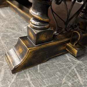 May include: Close-up of a dark wood table leg with a black and gold finish. The leg has a decorative scroll design and a square base.