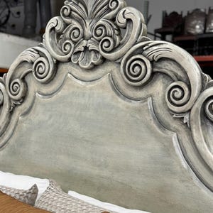 May include: Close-up of a gray-painted headboard with ornate, carved details. The headboard features a scrolling design with a central floral motif. The bed is partially visible with pillows and bedding in the foreground.
