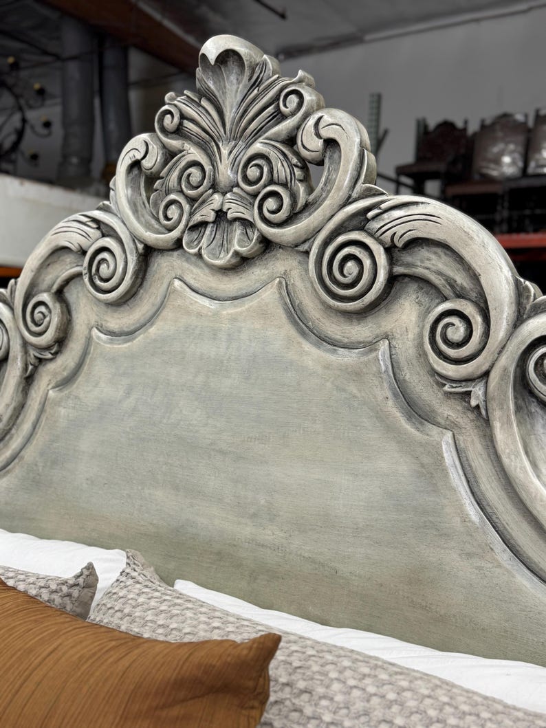 May include: Close-up of a gray-painted headboard with ornate, carved details. The headboard features a scrolling design with a central floral motif. The bed is partially visible with pillows and bedding in the foreground.