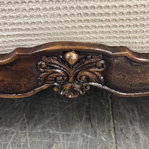 K&ouml;nnte beinhalten: Nahaufnahme eines Teils eines dunkelbraunen Holzm&ouml;bels mit kunstvollen Schnitzereien. Die Schnitzereien sind mittig angeordnet und weisen ein florales Design auf. Der Hintergrund ist ein hellbeige strukturiertes Gewebe.