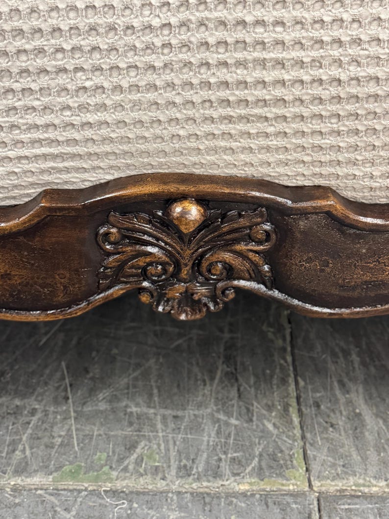 K&ouml;nnte beinhalten: Nahaufnahme eines Teils eines dunkelbraunen Holzm&ouml;bels mit kunstvollen Schnitzereien. Die Schnitzereien sind mittig angeordnet und weisen ein florales Design auf. Der Hintergrund ist ein hellbeige strukturiertes Gewebe.
