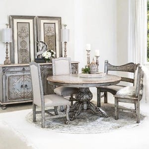 May include: A round, distressed grey wood dining table with a carved pedestal base. The table is surrounded by four chairs, including a bench with a curved back. The chairs are also distressed grey wood with upholstered seats. The room is decorated in a farmhouse style with a white rug and grey walls.