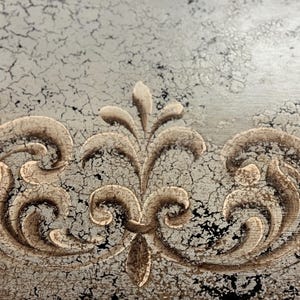 May include: A close-up of a decorative design on a distressed, silver-painted surface. The design features a central floral motif with swirling, scroll-like patterns on either side.