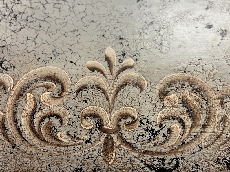 May include: A close-up of a decorative design on a distressed, silver-painted surface. The design features a central floral motif with swirling, scroll-like patterns on either side.