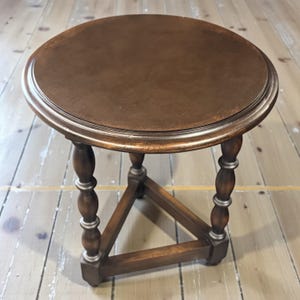 May include: A small, round, wooden side table with a dark brown tabletop and a silver trim. The table has three turned legs with silver accents and a triangular base for stability. The table is a classic piece of furniture.