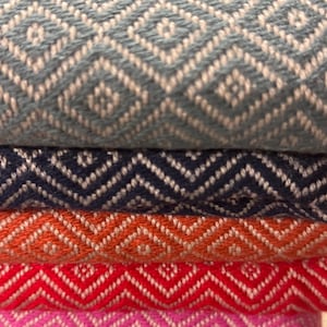May include: A stack of colorful woven textiles with geometric patterns. The top textile is a light blue with white diamond shapes. Below are navy, orange, red, and pink textiles, each with a zig-zag design. The fabrics appear soft and textured.