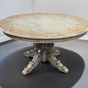 May include: A round, white, wooden table with a carved, ornate base and a distressed, light green top with a gold-coloured design.