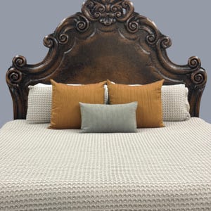 K&ouml;nnte beinhalten: Ein dunkelbraunes, verziertes Kopfende mit einem geschnitzten Design. Das Bett ist mit einer hellbeigen, strukturierten Decke und mehreren Kissen in Orange- und Graut&ouml;nen ausgestattet. Das Bett ist gemacht und bezugsfertig.