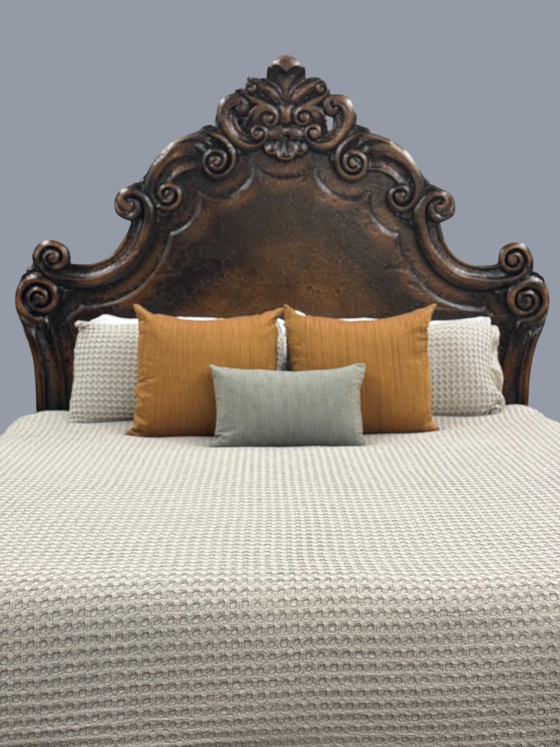 K&ouml;nnte beinhalten: Ein dunkelbraunes, verziertes Kopfende mit einem geschnitzten Design. Das Bett ist mit einer hellbeigen, strukturierten Decke und mehreren Kissen in Orange- und Graut&ouml;nen ausgestattet. Das Bett ist gemacht und bezugsfertig.