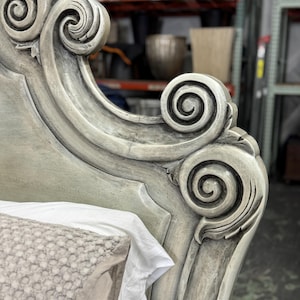 May include: Close-up of a distressed, ornate headboard with scroll detailing. The headboard is painted in a muted gray-green color, with visible texture and carved accents. A white pillow and sheet are partially visible.