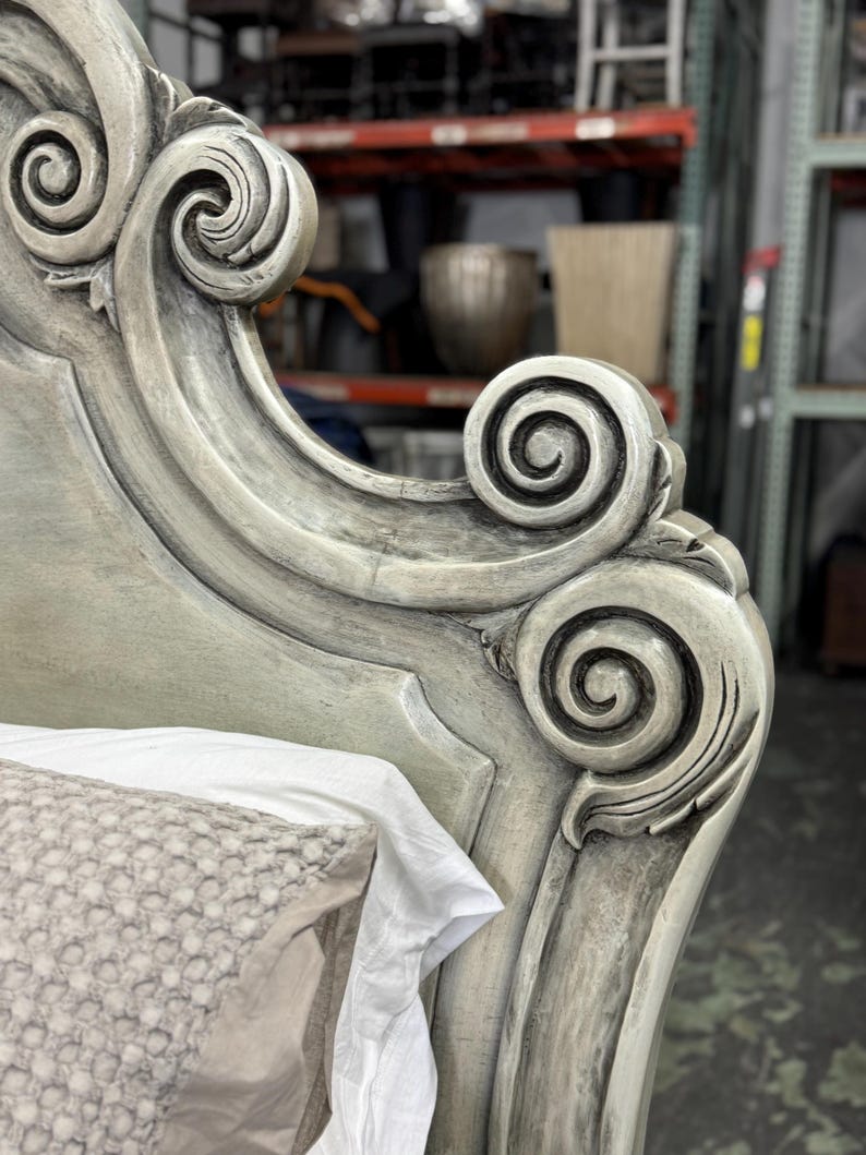 May include: Close-up of a distressed, ornate headboard with scroll detailing. The headboard is painted in a muted gray-green color, with visible texture and carved accents. A white pillow and sheet are partially visible.