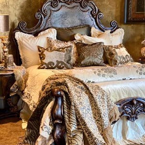 K&ouml;nnte beinhalten: Ein luxuri&ouml;ses Bett mit einem geschnitzten Kopfteil und aufwendigen Details. Das Bett ist mit Kissen und einer gemusterten Bettdecke geschm&uuml;ckt. Eine dekorative Decke mit Fransen ist &uuml;ber dem Fu&szlig;ende des Bettes drapiert. Die Farbpalette umfasst Gold, Braun und Creme.
