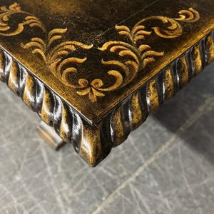 May include: Close-up of a dark brown table top with a gold leaf design. The table top has a rope-like edge.