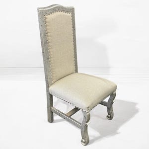 May include: A grey upholstered dining chair with a high back and a carved wooden frame. The chair has a tufted back and a nailhead trim.