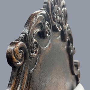 K&ouml;nnte beinhalten: Nahaufnahme einer dunkelbraunen, verzierten Holzlehne. Das Design weist aufwendige Schn&ouml;rkel und florale Motive mit einem Used-Look auf. Die Stuhllehne hat eine geschwungene Oberseite und eine rechteckige Form.
