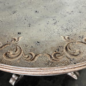May include: A round table top with a distressed grey paint finish and a decorative gold-coloured scroll design around the edge.
