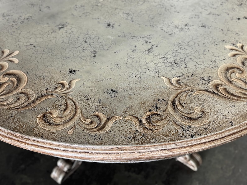 May include: A round table top with a distressed grey paint finish and a decorative gold-coloured scroll design around the edge.