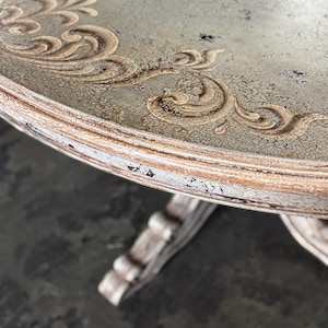 May include: A round, distressed, light green table top with a decorative, gold-painted, scroll design around the edge. The table has a distressed, white-painted base with ornate details.