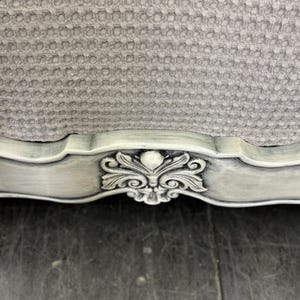 May include: Close-up of a section of a light gray upholstered chair. The chair has a detailed, ornate, carved wooden frame painted in a distressed, off-white finish. The upholstery has a textured, honeycomb pattern.