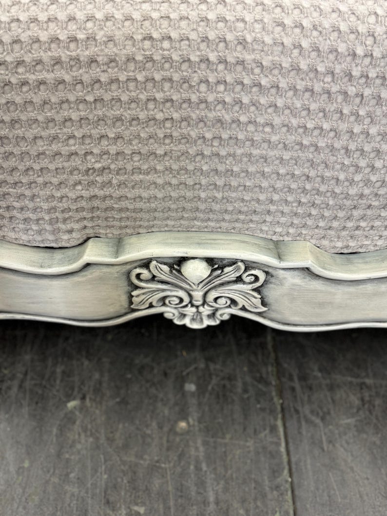 May include: Close-up of a section of a light gray upholstered chair. The chair has a detailed, ornate, carved wooden frame painted in a distressed, off-white finish. The upholstery has a textured, honeycomb pattern.