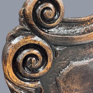 K&ouml;nnte beinhalten: Nahaufnahme eines dekorativen M&ouml;beldetails in Bronzefarbe. Das Design zeigt eine scrollartige Verzierung mit strukturierter Oberfl&auml;che und dunklen Akzenten. Das St&uuml;ck hat eine Vintage- oder Antik-&Auml;sthetik.