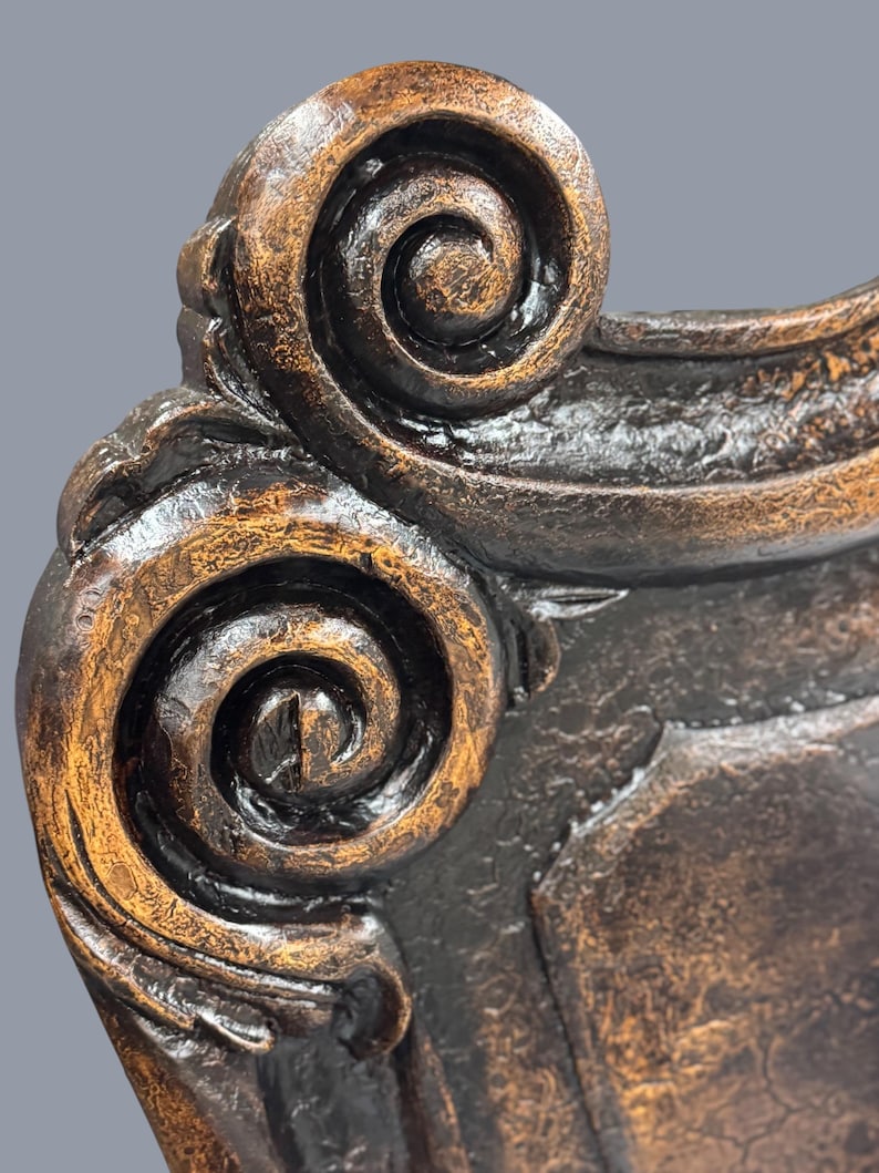 K&ouml;nnte beinhalten: Nahaufnahme eines dekorativen M&ouml;beldetails in Bronzefarbe. Das Design zeigt eine scrollartige Verzierung mit strukturierter Oberfl&auml;che und dunklen Akzenten. Das St&uuml;ck hat eine Vintage- oder Antik-&Auml;sthetik.